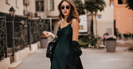 Fashionable pale brunette in long green dress, black jacket and sunglasses, standing on street duri
